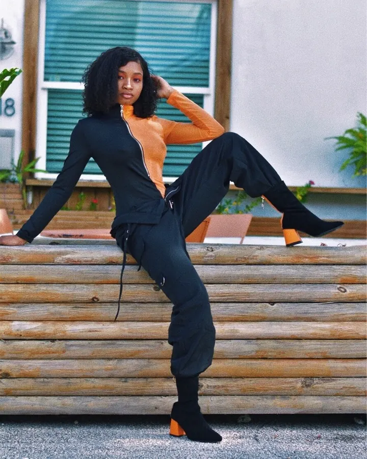 Femme en tenue noire et orange, assise sur un banc en bois, regardant l'appareil photo