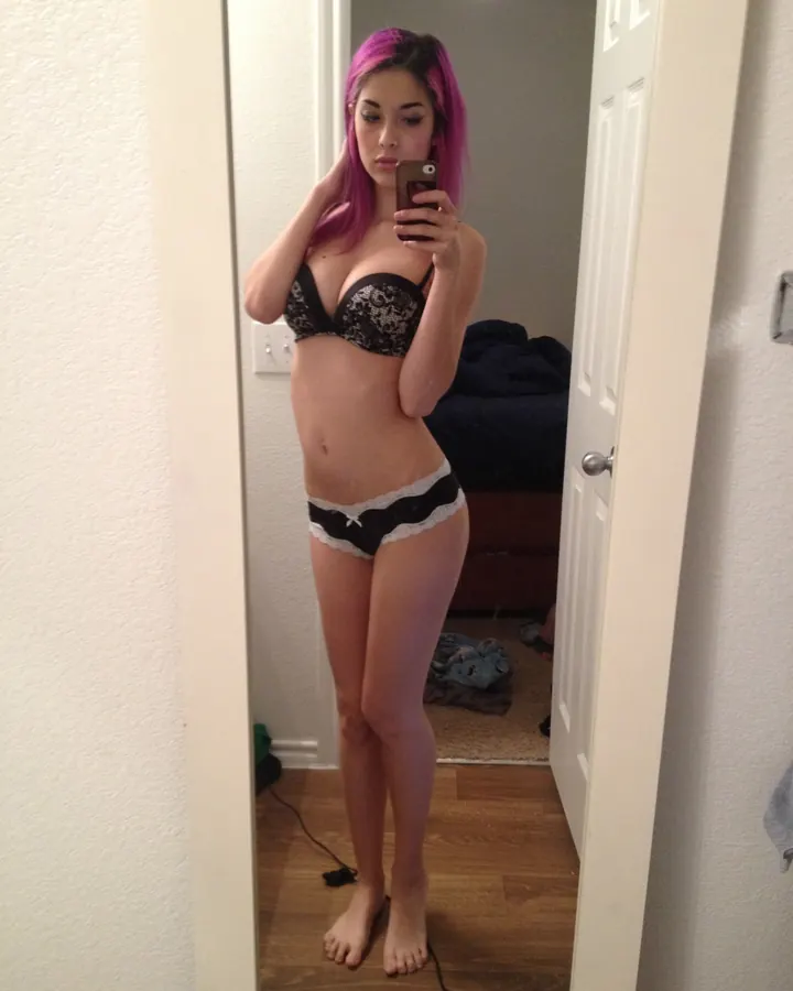 Femme aux cheveux bruns foncés, vêtue d’un haut noir et d’un corset noir, prenant un selfie en tenant un téléphone, regardant vers l'appareil.