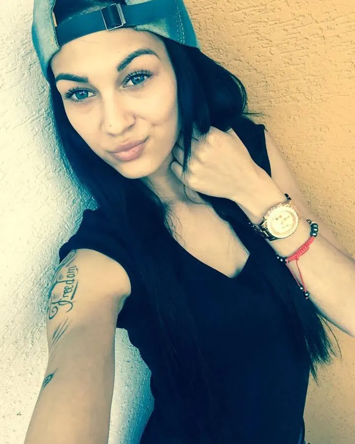Femme latino aux yeux bleus, coiffée d'une casquette renversée, portant un haut noir, un tatouage visible sur le bras et une montre dorée.