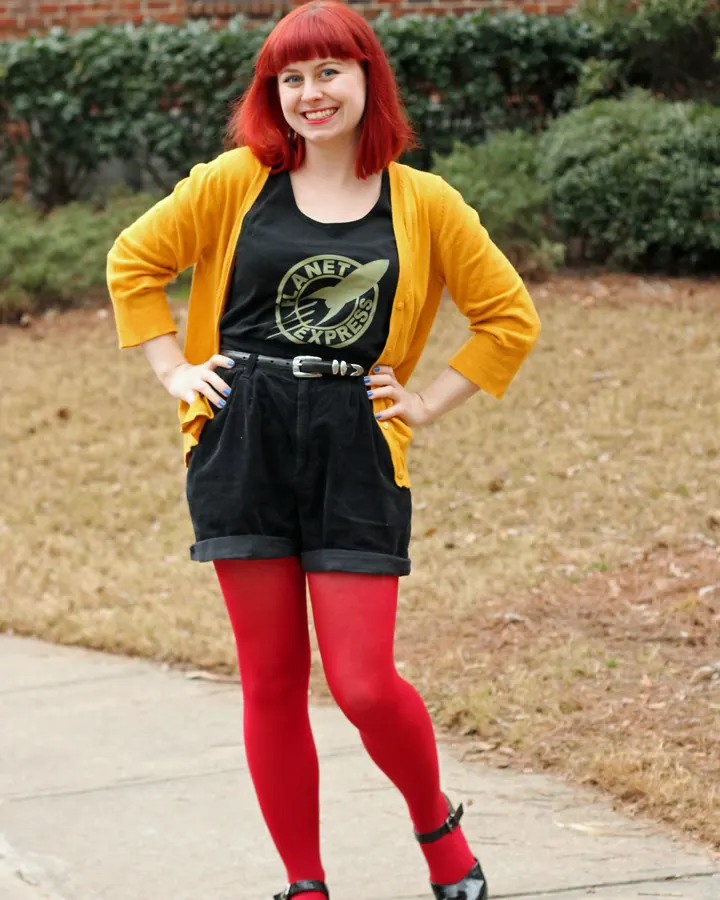 Femme européenne aux cheveux roux, portant un cardigan jaune, un t-shirt noir avec un logo, un short noir, des collants rouges et des chaussures noires, souriante.