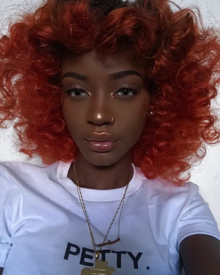 Femme africaine aux cheveux roux bouclés, portant un t-shirt blanc avec l'inscription 'PETTY.' et un collier doré, regard fixe.