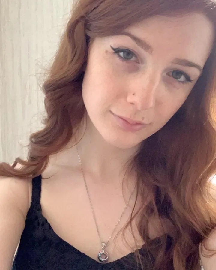 Femme aux cheveux roux en haut noir, portant un collier, regardant vers l'appareil photo.