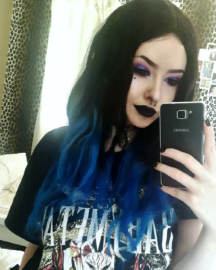 Femme aux cheveux noirs, maquillage sombre, cheveux teints en bleu, prenant un selfie dans une chambre, vêtue d'un t-shirt graphique.