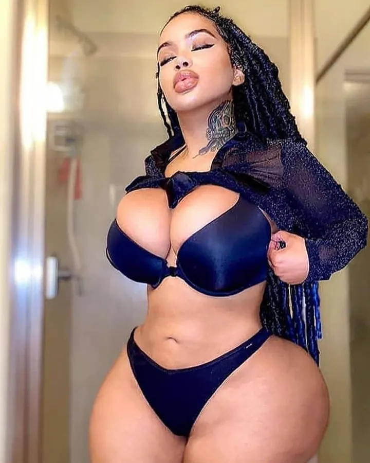 Femme africaine aux cheveux longs tressés, vêtue d'un soutien-gorge bleu et d'une robe transparente, pose dans un couloir.
