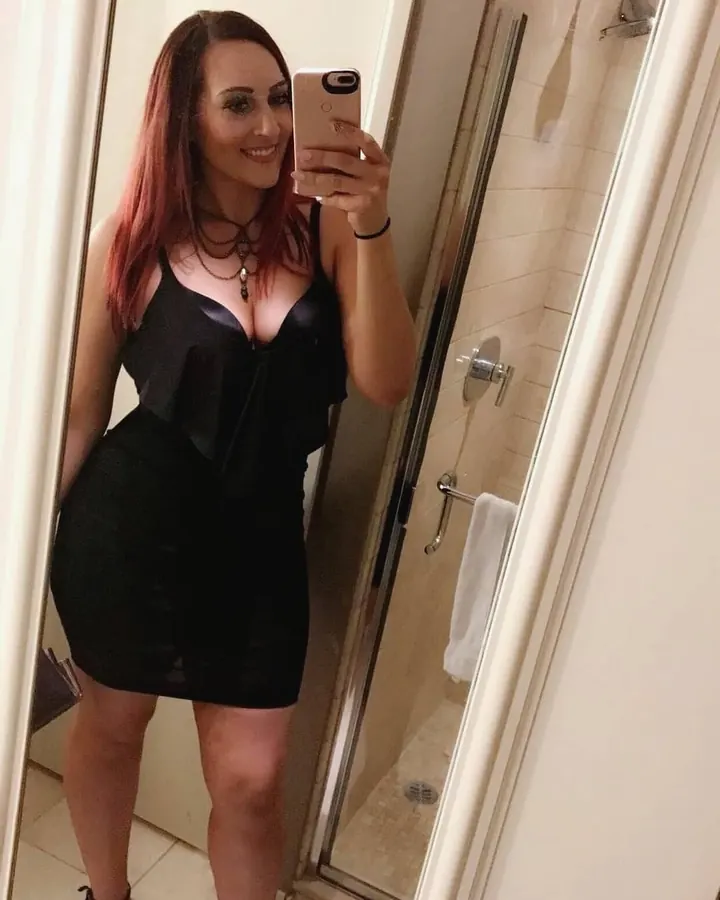 Femme aux cheveux roux, en robe noire courte, souriant dans une salle de bain, prenant un selfie.