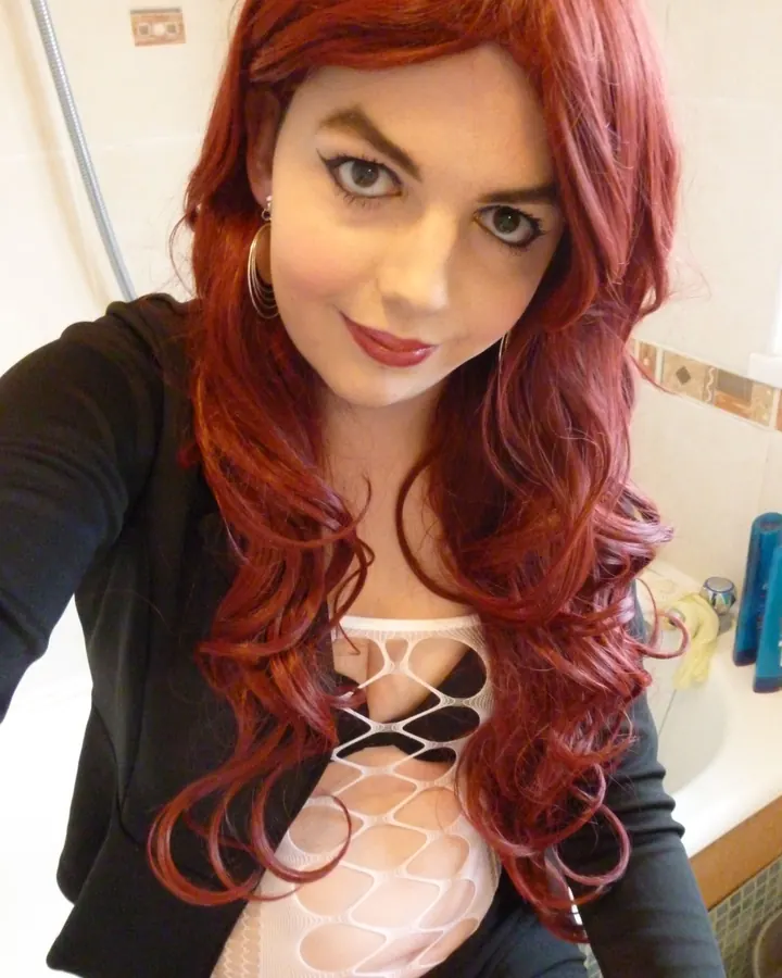 Femme aux cheveux rouges en haut blanc ajouré, posant dans une salle de bain.