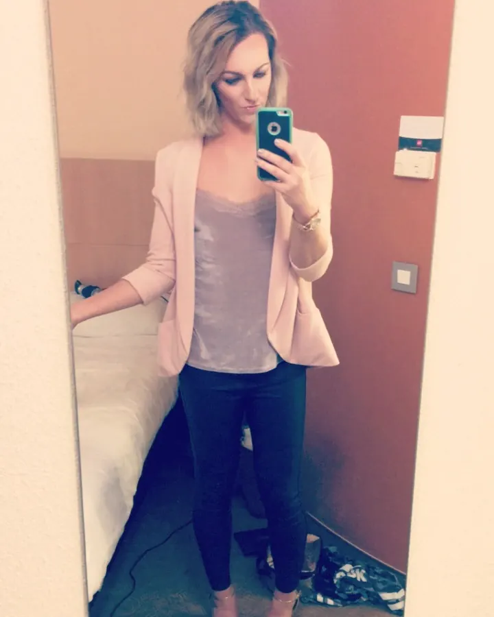 Femme blonde en veste rose et jeans, prenant un selfie devant un miroir.
